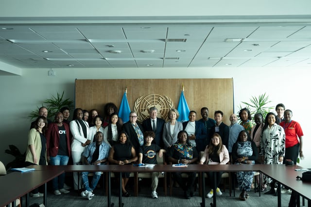 Advocacy Week participants pose for a group photo in a meeting room at United Nations headquarters, in front of the UN emblem and two UN flags.