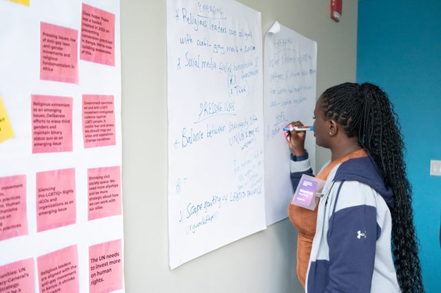 A workshop participant writes on a flip chart beside a wall of pink sticky notes.