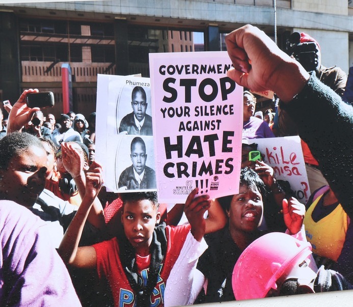 Protesters hold a sign that reads "Government, stop your silence against hate crimes" beside photos of a young person, in South Africa
