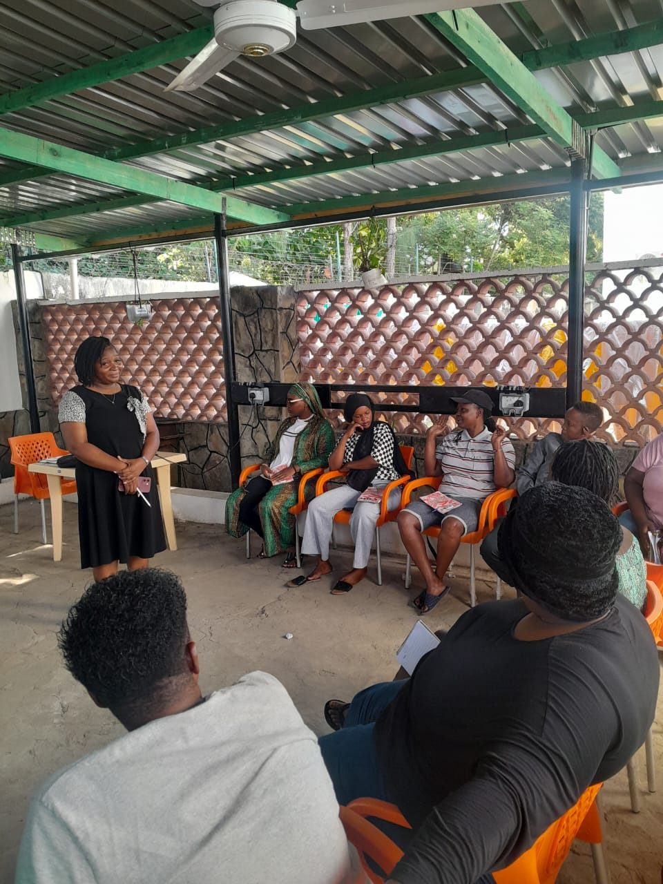 A trainer speaks to a seated group of participants in an open-sided hall during a childcare skills session