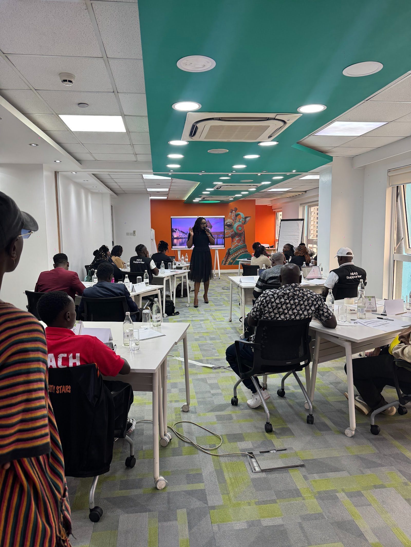 Participants seated at tables in a bright training room listen to a facilitator